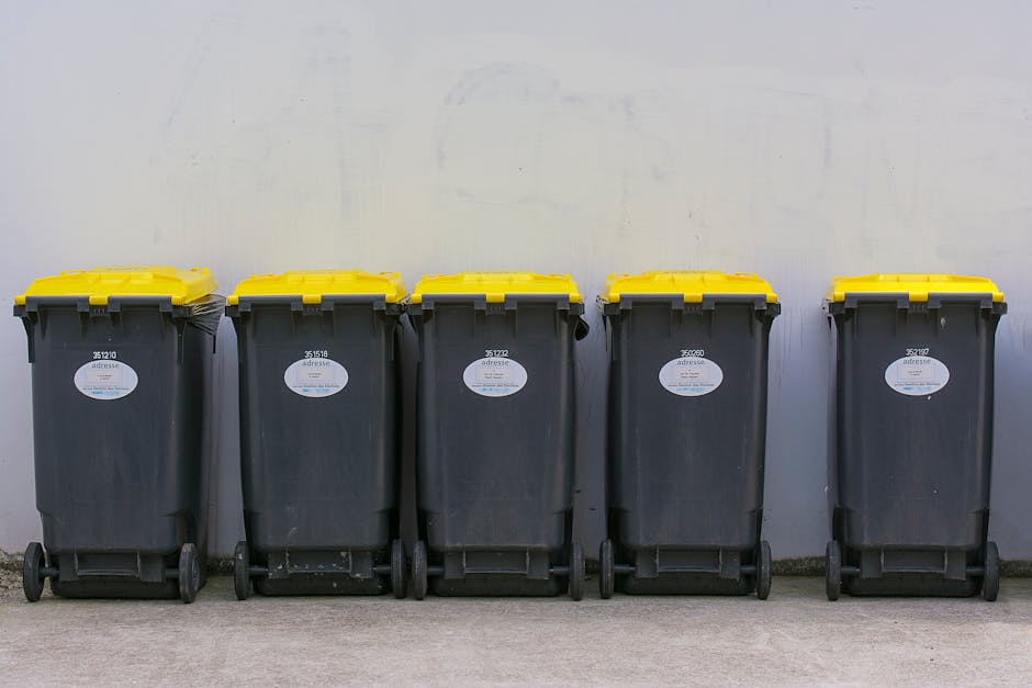 A row of five large wheelie bins made of durable black plastic with yellow lids, positioned on a paved surface against a plain light-colored wall. The bins are evenly spaced and aligned horizontally, with each bin featuring a white label with black text and barcodes on the front. The environment appears to be an outdoor area, likely a residential or commercial setting where waste collection occurs, supported by the presence of a concrete or paved ground surface. The yellow lids are securely closed, indicating readiness for waste disposal or collection, which is typical in private waste handling or alternative rubbish removal services like those provided by Rubbish Removal Wimbledon. The scene suggests a setting for independent rubbish collection, emphasizing the importance of organized waste management and disposal options outside standard municipal services, aligning with the theme of efficient rubbish removal for flats or properties.