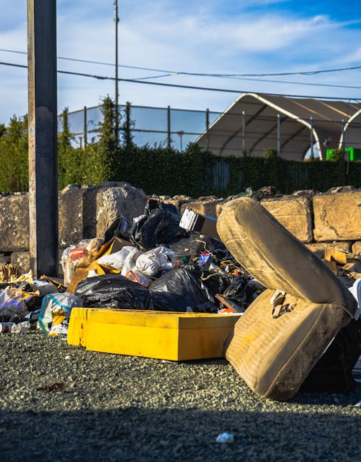 A roadside pile of discarded rubbish includes black plastic garbage bags, a yellow plastic container, and an old, worn car tyre lying on its side. The waste is situated at the base of a stone wall made from rough, uneven blocks. In the background, a black metal fence runs parallel to the stone wall, with green foliage and small trees visible behind it. Overhead, part of a curved fabric-covered structure, possibly a tennis or sports court shelter, is visible, along with power lines stretching across a bright sky with scattered clouds. The scene suggests an informal or unscheduled rubbish dump, indicative of private waste disposal or collection services, such as those offered by Rubbish Removal Wimbledon, to manage non-standard waste on-site. The environment appears outdoors on a pavement or driveway area, with natural daylight illuminating the waste and surroundings, emphasizing the need for proper rubbish collection to maintain cleanliness and environmental standards.