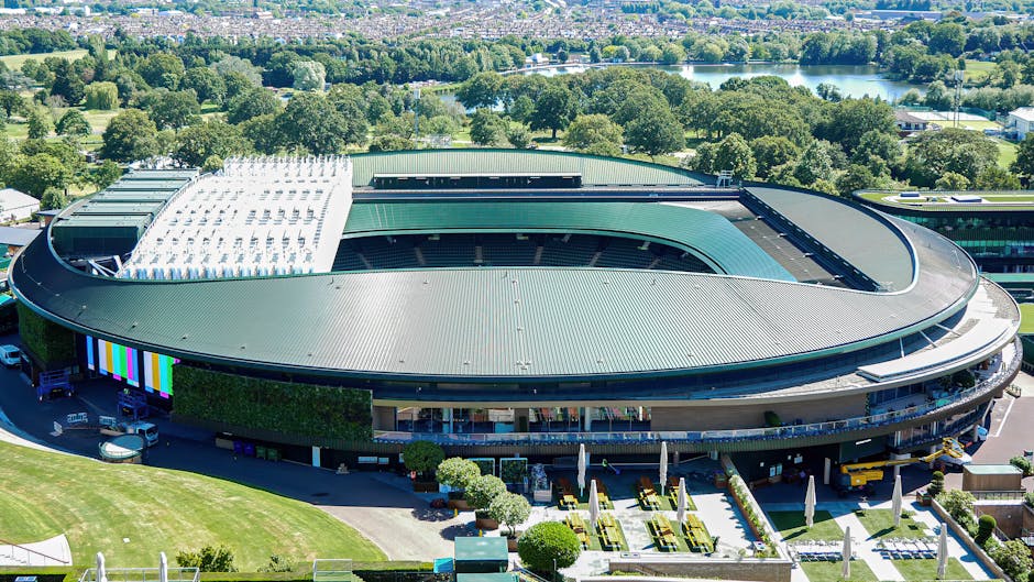 An aerial view of a large, modern sports stadium with an oval-shaped green metal roof, featuring a central open area and a section on one side covered with white solar panels. The stadium's exterior walls are dark, with some landscaped areas and outdoor seating visible along the perimeter, including white umbrellas and neatly trimmed bushes. Surrounding the stadium is a well-maintained lawn and paved areas, with vehicles parked nearby. In the background, there are numerous trees, a body of water, and a distant view of greenery, indicating a park-like setting. The scene is illuminated by daylight, with clear weather providing bright natural light, which highlights the architectural details and the lush environment around the stadium. This image, associated with waste removal and site clearance services, suggests the importance of maintaining an environmentally conscious approach when handling large-scale facilities or renovation projects, aligning with the principles of private waste disposal and alternative rubbish collection methods.