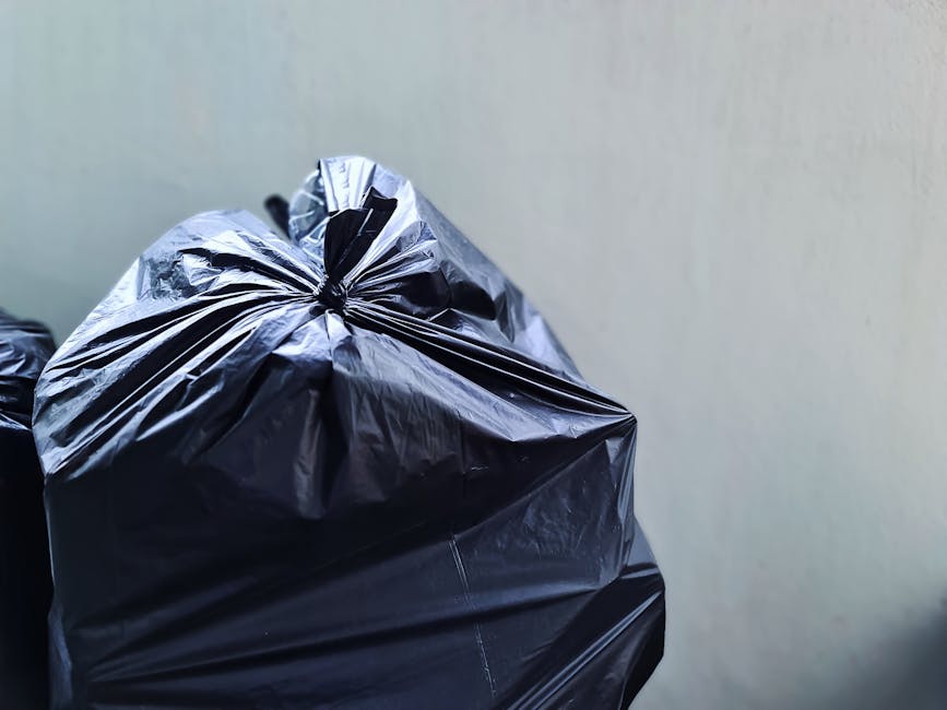 Close-up view of a large black plastic rubbish bag tightly knotted at the top, placed on a flat surface against a plain light grey wall. The bag appears to be made of glossy, durable plastic with visible creases and slight reflections due to the lighting. In the background, additional black bags are partially visible, suggesting a collection of waste awaiting removal. The scene is set indoors or in a sheltered area, indicative of a private or commercial waste collection point. The environment is minimally detailed, focusing on the bag's material and shape, which is common in refuse disposal contexts. This image visually supports private rubbish handling services, such as waste collection or waste management solutions offered by Rubbish Removal Wimbledon, highlighting the need for efficient rubbish disposal methods for timely and proper waste removal.