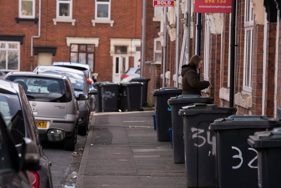A residential street scene shows a row of black wheelie bins placed along the sidewalk outside brick-built terrace houses, with some bins labeled with painted white numbers. The bins are positioned close to each other, with the majority facing the street and some slightly angled towards the façade of the buildings. The houses have red brick facades, white framed windows, and stone window sills, and some feature small external doors or porches. A woman, dressed in a brown coat, is seen standing near one of the bins, possibly about to open or close it. Parked cars are lined up along the edge of the road, including a small silver hatchback nearby in the foreground, with more vehicles further down the street, extending into the background. The environment appears typical of an urban residential area with a narrow road and limited space for refuse collection, which may suggest the need for independent waste handling such as bin collection services. This scene aligns with private rubbish removal efforts, showcasing onsite waste management outside a row of homes, with the focus on the bins and street context, suitable for content related to rubbish removal in urban settings.